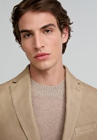 Blazer de algodón beige con un bolsillo en el pecho, superpuesto a un suéter de punto acanalado marrón claro. Textura suave, diseño estructurado.