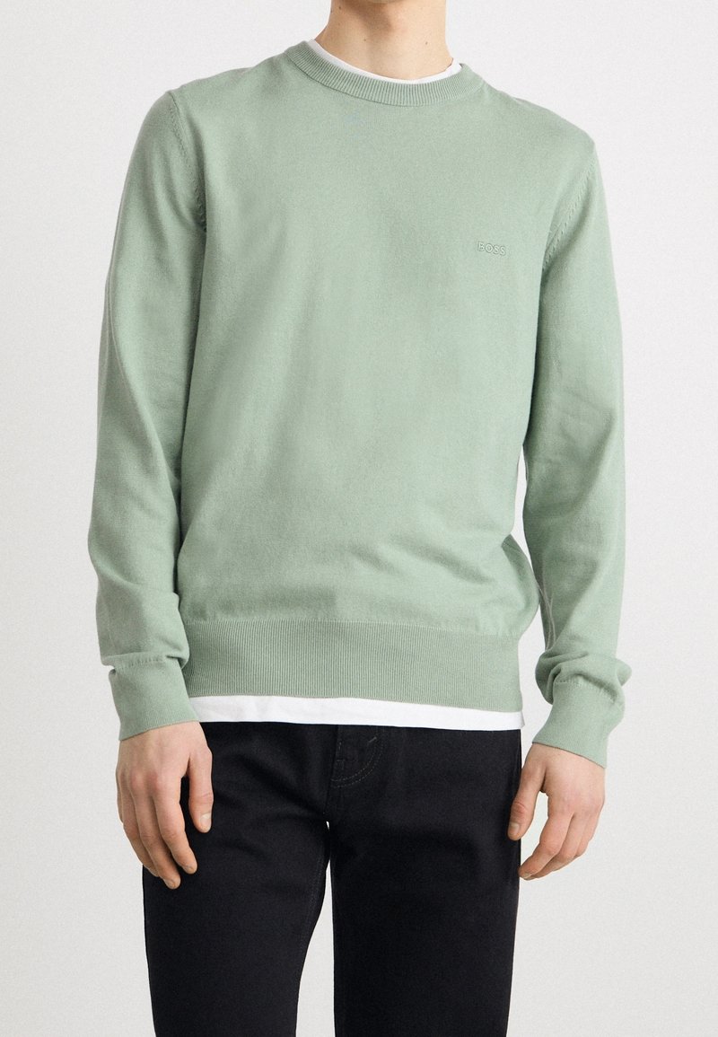 Ljusgrön crew-neck tröja i mjukt tyg, med ribbade muddar och nederkant, lagd över en vit skjorta. Svarta byxor fulländar looken.