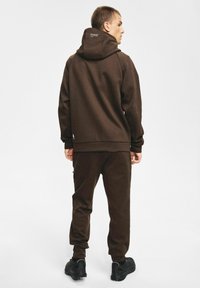 Brązowa bluza z kapturem i spodnie dresowe wykonane z miękkiego materiału. Bluza ma kaptur z logo, rękawy i nogawki zakończone ściągaczami. Buty są czarne z teksturowaną podeszwą.