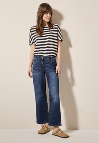 Gestreepte gebreide top in crème en marineblauw, gecombineerd met een wijdvallende spijkerbroek van donker denim en taupe instapschoenen, staande tegen een neutrale achtergrond.