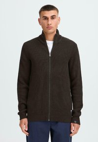 Dunkelbrauner gerippter Strickzipper mit hohem Kragen und langen Ärmeln, mit einem kleinen Logo-Detail am Bündchen. Einfarbig.