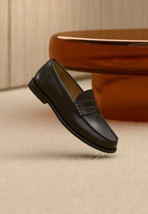 Scarpa mocassino in pelle nera sospesa sopra un tappeto beige con mobili moderni color ambra sullo sfondo.