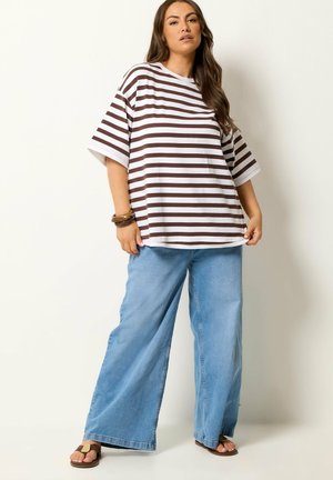 Kvinde iført oversized brun og hvid stribet t-shirt, lyseblå jeans med vide ben, sandaler og brune armbånd, står mod en ensfarvet baggrund.