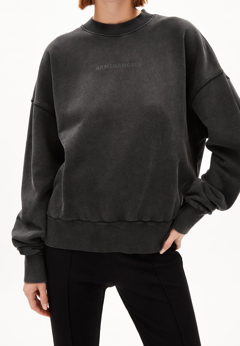 Femme portant un sweat-shirt noir délavé avec le texte "ARMEDANGELS" sur la poitrine et un pantalon noir, main reposant sur la hanche.