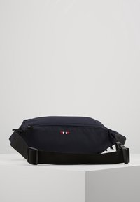 Marsupio blu navy con tasca frontale con cerniera, piccolo logo a strisce rosse, bianche e blu, e tracolla nera regolabile con fibbia in plastica.