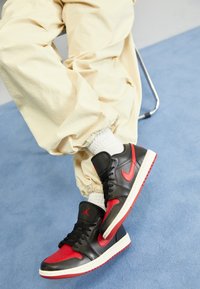 Osoba na kovové židli, oblečená v černých a červených teniskách Nike Air Jordan s bílými ponožkami a béžovými kalhotami, sedí na modré podlaze.