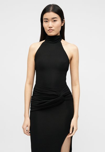 Γυναίκα με μαύρο αμάνικο φόρεμα halter neck με ψηλή σκίσιμο και λεπτομέρεια συγκέντρωσης ύφασματος στη μέση, που στέκεται απέναντι σε απλό φόντο.