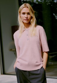 Pull en tricot rose clair à manches courtes avec un ourlet et un col côtelés ; présente trois boutons décoratifs. Associé à un pantalon sombre.
