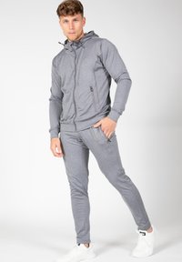 Grijze sportjack met rits en capuchon, gecombineerd met bijpassende tapered broek. Gladde textuur, met ritszakken en een aangesloten ontwerp.