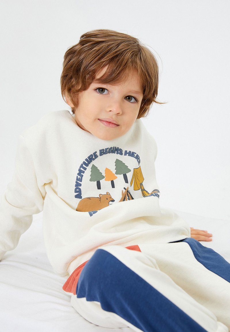 Sudadera de color crema con un gráfico colorido al aire libre que incluye árboles, un oso y una tienda de campaña. Combinada con pantalones de chándal azul y blanco y un acento rojo.