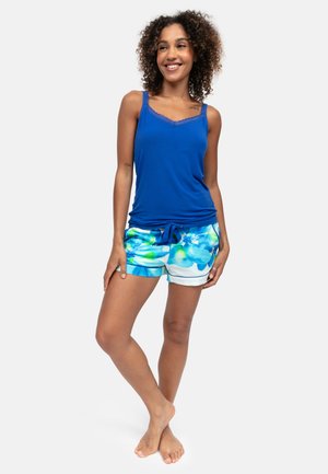 Vrouw staat op blote voeten, draagt een blauw mouwloos topje en blauwe korte broek met bloemenprint, glimlacht met krullend haar en een ontspannen houding.