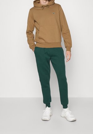 Felpa con cappuccio marrone dotata di tasca frontale, abbinata a pantaloni sportivi a gamba affusolata verdi scuri. Sneakers bianche completano l'outfit.