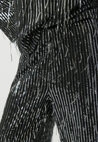 Zwarte stof met metallic zilveren strepen en pailletten. De textuur lijkt glanzend met losse draadjes en franjerandjes langs de randen.