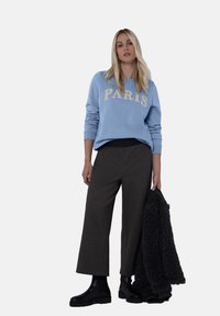 Jasnoniebieska bluza z napisem „PARIS” w beżowych literach, zestawiona z szerokimi spodniami w kolorze węgla drzewnego i czarnymi botkami, trzyma ciemną kurtkę imitującą futro.