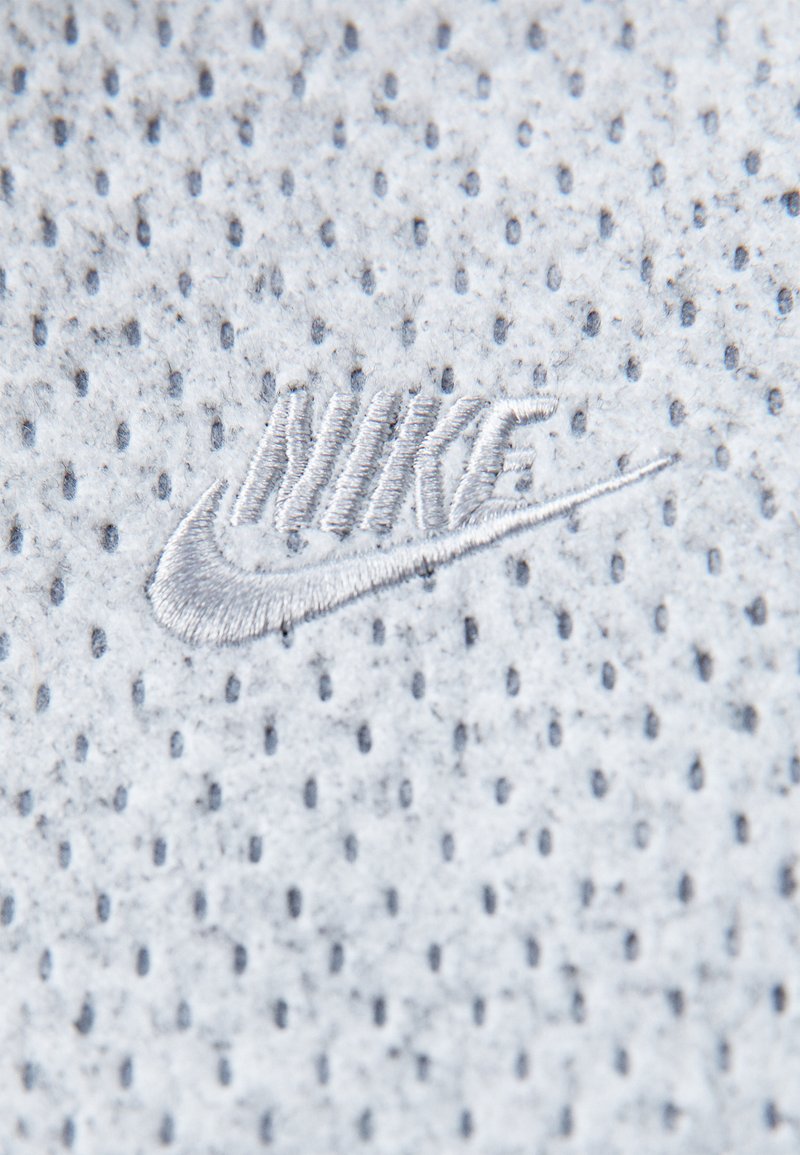 Hellgrauer Stoff mit durchbrochener Textur, der ein weiß gesticktes Nike-Logo zeigt und eine glatte Oberfläche sowie subtile Details aufweist.