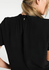 Femme aux cheveux blonds attachés portant un haut noir avec une petite fermeture à bouton à l'arrière du col.