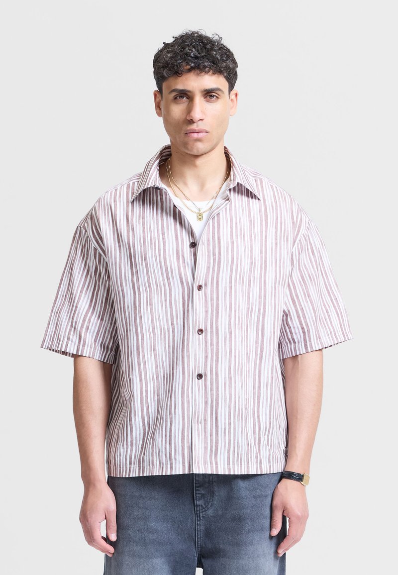 Homme portant une chemise à manches courtes à rayures verticales marron et blanches, des colliers en or superposés, un jean foncé et une montre-bracelet, debout en position neutre.