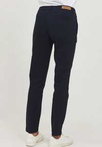 Marinblå bomullsbyxor med rak passform, med en bakficka och en beige logopatch vid midjan, i kombination med vita sneakers.