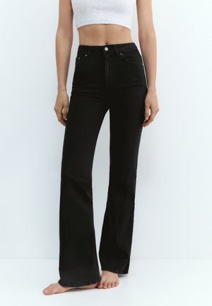 Žena u crnim trapericama visokog struka sa širokim nogavicama i bijelom kratkom majicom, stoji bosa na jednostavnoj bijeloj pozadini.