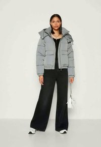Grey puffer jacket with a hood, featuring a boxy design, multiple pockets, and a smooth texture. Paired with black wide-leg pants and white sneakers.