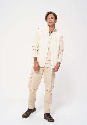 Kremowa polarowa kamizelka na beżowym swetrze w prążki, w połączeniu z beżowymi spodniami cargo i brązowymi zausznikami, na tle jednolitego tła.