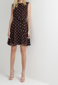 Ruskeakuvioinen polka dot -mekko, jossa on istuva yläosa, levenevä helma ja vyötärönauha, valmistettu kevyestä kankaasta. Yhdistetty vaaleisiin korkokenkiin.