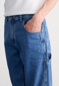 Modré džíny z denimu s uvolněným střihem, boční kapsou a viditelným knoflíkem na pásu. Látka má jemnou texturu.