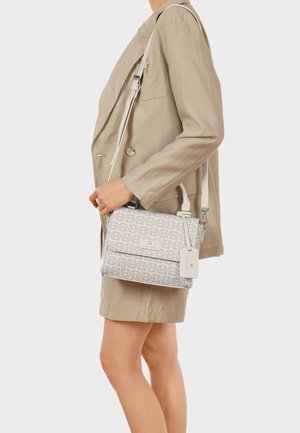 Donna che indossa un blazer e una gonna beige, con una borsa bianca decorata con tracolla regolabile e un piccolo tag per bagaglio.