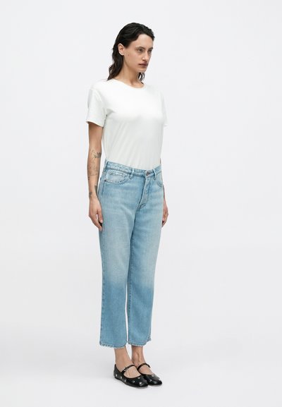 Λευκό κοντομάνικο μπλουζάκι, ανοιχτό μπλε τζιν παντελόνι ίσιας γραμμής με φθαρμένες λεπτομέρειες και μαύρα επίπεδα παπούτσια με κυκλικές διακοσμήσεις.