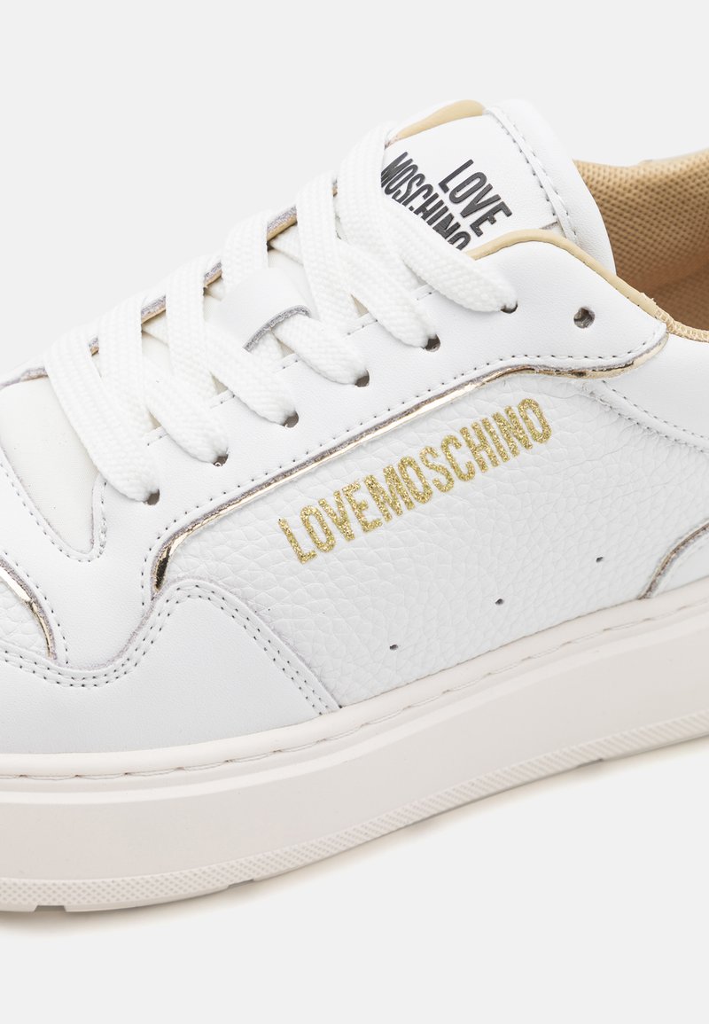 Sneakers bianchi in pelle testurizzata, con logo in accento dorato, lacci piatti e una robusta suola bianca. Caratterizzati da un design minimal e cuciture rinforzate.