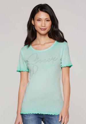 Woman with long dark hair wearing a light green short-sleeve shirt with scalloped edges and subtle glittery text on the front.