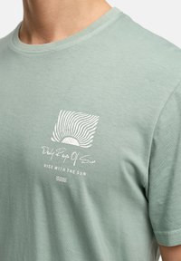T-shirt en coton vert clair avec un col rond, des manches courtes et un design graphique à l'avant représentant des rayons de soleil et du texte en blanc.