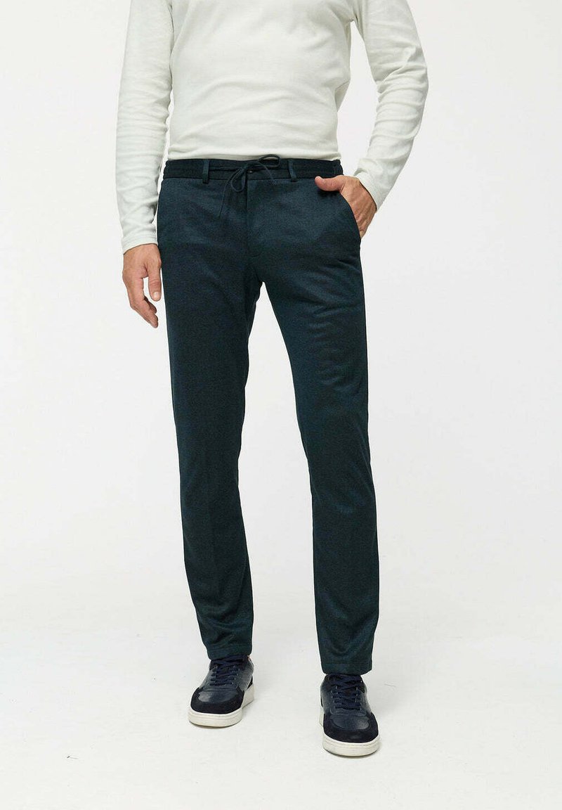 Dunkelgrüne Hose mit einem Kordelzugbund, taillierter Passform und glatter Textur. Kombiniert mit einem weißen Langarmshirt und marineblauen Sneakers.