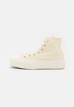 Baskets montantes en toile beige avec semelle en caoutchouc blanche et lacets blancs, montrées de profil sur un fond clair uni.