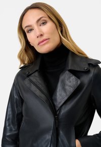 Giacca da biker in pelle nera con colletto strutturato, chiusura a zip e texture liscia. Presenta bottoni a pressione e design aderente.