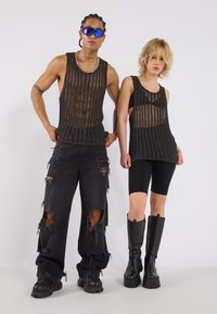 Zwei Personen tragen schwarze, übergroße Strick-Tanktops mit offener Webung. Die eine kombiniert es mit zerrissenen schwarzen Jeans, die andere mit schwarzen Radshorts und kniehohen Stiefeln.