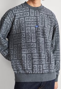 Sudadera gris con un patrón repetido del logo "HUGO", cuello redondo y puños y dobladillo de canalé. Textura suave, ajuste casual.
