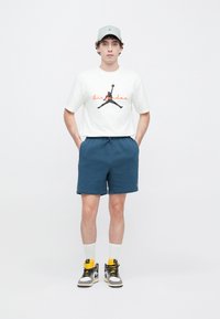 T-shirt blanc avec logo Air Jordan noir et texte orange, accompagné d'un short bleu marine, de chaussettes blanches et de baskets noires et blanches.