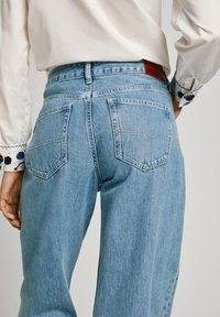 Ljusblå denimjeans med hög midja, utrustade med två bakfickor och en brun läderlapp vid midjan, visade bakifrån.