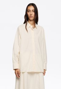 Camisa oversize beige claro confeccionada en una tela suave, con botonadura frontal, cuello puntiagudo y mangas largas. Textura lisa.