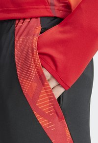 Rode sporttop met grafische patronen en zwarte shorts met een ingestikte ritszak. Glad materiaal, aansluitend ontwerp en contrasterende kleuren.