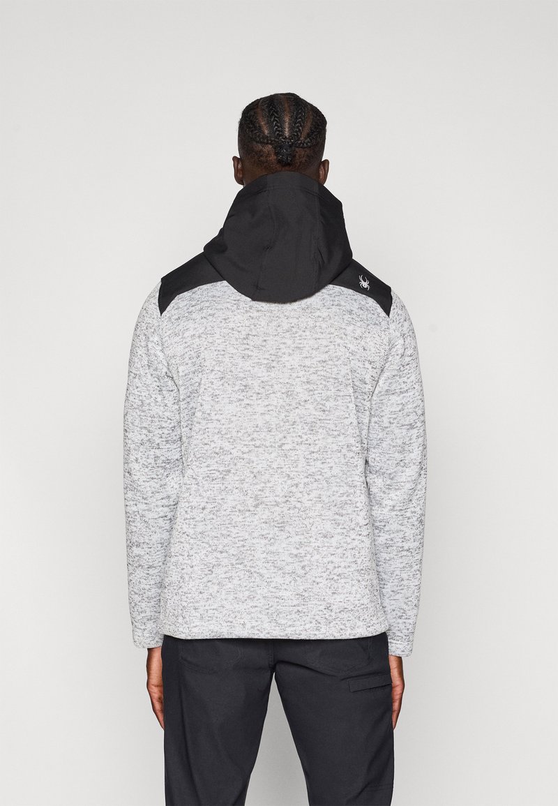 Veste grise et noire avec un tricot texturé, dotée d'un col montant et d'une grande capuche, conçue pour la chaleur et le confort.