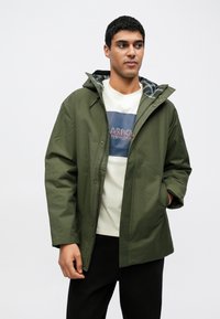 Casaco impermeável verde-oliva com capuz, com forro xadrez, fechos de botões e corte descontraído. Usado sobre uma t-shirt creme.