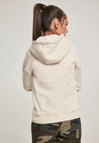 Krémová mikina s kapucňou s jemnou textúrou, voľným strihom, predným klokaním vreckom a kapucňou so sťahovacou šnúrkou, v kombinácii s maskáčovými nohavicami.
