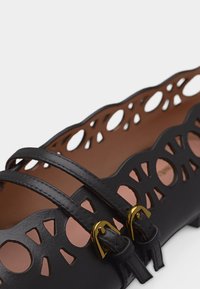 Μαύρο πλατυπόδεμο παπούτσι με διακοσμητικές περικοπές κατά μήκος των άκρων και δύο λεπτές λουρίδες που στερεώνονται με μικρές χρυσές αγκράφες.