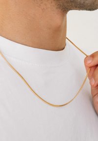 Guld kæde med en snoet design, hvilende på en hvid t-shirt, holdt af en hånd nær udskæringen. Glat, skinnende tekstur.