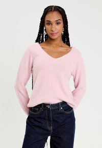 Vaaleanpunainen, pörröinen V-kaula-aukkoinen neulepusero pitkillä hihoilla, yhdistetty tummansinisiin farkkuihin. Neuleen pinta on pehmeä ja istuvuus väljähkö.