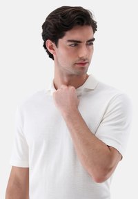 Weißes Poloshirt mit kurzen Ärmeln aus weichem Stoff, das einen klassischen Kragen aufweist, eine glatte Textur und ein minimalistisches Design hat.