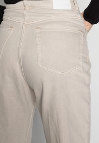 Pantalones de mezclilla beige con corte recto, que cuentan con bolsillos traseros y sutiles detalles de costura. Textura suave con un acabado liso.