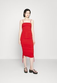 Vestido justo vermelho com alças finas; textura franzida nas laterais; comprimento até ao joelho com laços ajustáveis nas laterais; modelo a usar sandálias plataforma pretas.